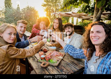 Gruppe von multirassischen Freunden, die Selfies machen und mit Weingläsern zum Abendessen auf der Terrasse feiern - Jugendliche essen und trinken Rotwein Stockfoto