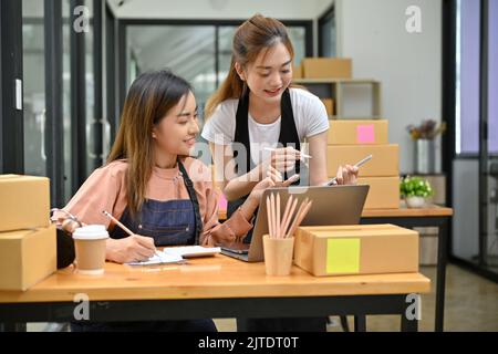 Zwei attraktive junge asiatische Freundinnen Online-Shop-Besitzer arbeiten zusammen in der Lagerhalle, Versandkartons vorbereiten und Bestellungen überprüfen. E-Comer Stockfoto