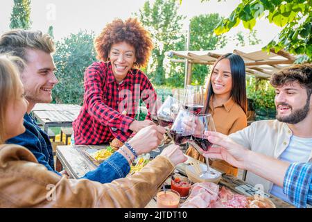 Gruppe von multirassischen Freunden, die Selfies machen und mit Weingläsern zum Abendessen auf der Terrasse feiern - Jugendliche essen und trinken Rotwein Stockfoto