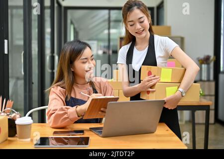 Zwei junge asiatische Freundinnen im Online-Shop arbeiten zusammen im Lagerraum, bereiten Versandkartons vor und überprüfen Bestellungen. Stockfoto