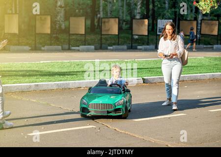 Mama steuert Kinderwagen mit Fernbedienung beim Spaziergang mit Kleinkind Kind im öffentlichen Park. Kleinkind Junge sitzt im grünen Auto halten Lenkrad zur Seite schauen Stockfoto