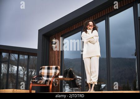 Lockige junge Frau, die auf der Terrasse des modernen Scheunenhauses in den Bergen ruht. Glückliche weibliche Touristen genießen in neue Hütte im Winter. Stockfoto