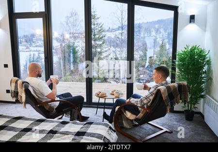 Zwei junge Männer genießen den Blick auf die winterliche Berglandschaft durch Panoramafenster in einem modernen Scheunenhaus. Glückliche männliche Touristen sitzen auf einem Stuhl und halten eine Tasse Tee in der neuen Hütte. Stockfoto