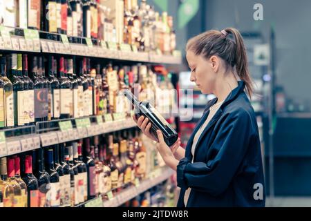 Eine Frau in einem Weinladen wählt, kauft Wein, liest das Etikett. Stockfoto