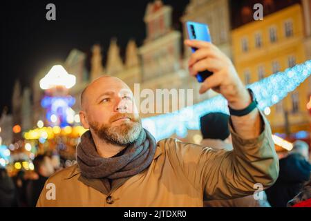 Ein Erwachsener fotografiert mit seinem Smartphone auf einem Weihnachtsmarkt in der Stadt. Der Mann nimmt Selfie mit dem Smartphone auf Stockfoto