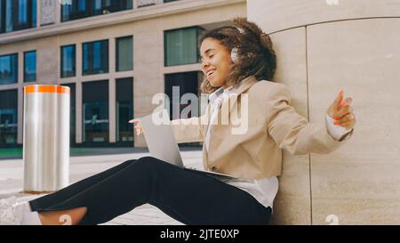 Afroamerikanische Frau mit weißen Kopfhörern hört Lieblingsmusik mit Vergnügen. Junge Dame tanzt, lehnt sich an die Säule und schaut auf den Laptop Stockfoto