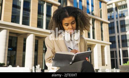 Afroamerikanische Frau schreibt Pläne in Papiernotizbuch mit nachdenklichen Ausdruck. Junge Geschäftsfrau denkt über zukünftige Start-ups nach, die sich Notizen durch Bürogebäude machen Stockfoto