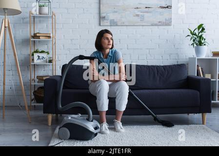 Junge Frau sitzt auf der Couch in der Nähe des Staubsaugers im Wohnzimmer Stockfoto