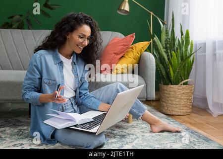 Junge schöne hispanische Studentin, die zu Hause aus der Ferne online studiert, Vortragsnotizen auf dem Boden sitzend im Wohnzimmer mit grüner Wand, Frau, die Kurse macht, die online studiert Stockfoto