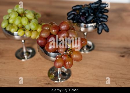 Ein Bund blauer, grüner und roter Trauben in Metallschüsseln auf einem Holztisch. Essen, gesunde Ernährung. Weicher, selektiver Fokus. Stockfoto