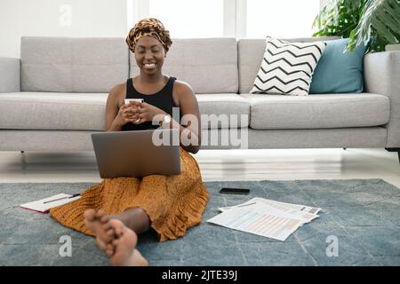 Schöne schwarze afrikanische Frau, die in der Lounge auf dem Boden sitzt, mit einem Laptop zu Hause mit Kaffee in der Hand und einem traditionellen Kopftuch Stockfoto