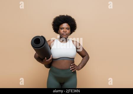 afroamerikanische plus size Frau in Crop top hält Fitness-Matte und stehend mit Hand auf Hüfte isoliert auf beige, Stock Bild Stockfoto