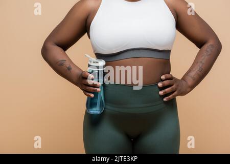 Beschnittene Ansicht der tätowierten afroamerikanischen plus size Frau in crop top hält Sportflasche mit Wasser isoliert auf beige, Stock Bild Stockfoto