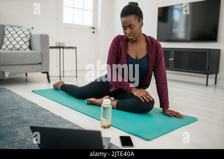 Schwarze Afrikanerin, die sich online auf dem Laptop zu Hause auf der Yogamatte im Wohnzimmer ausdehnt Stockfoto