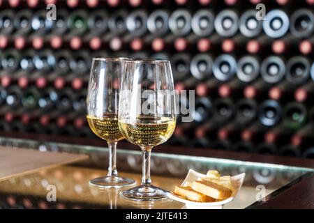 Weinverkostung in der Weinkellerei - 2 Gläser Weißwein mit Toast und Käse mit Hunderten von Weinflaschen dahinter, Lanzarote, Spanien Stockfoto