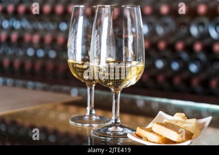 Weinverkostung in der Weinkellerei - 2 Gläser Weißwein mit Toast und Käse mit Hunderten von Weinflaschen dahinter, Lanzarote, Spanien Stockfoto