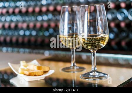 Weinverkostung in der Weinkellerei - 2 Gläser Weißwein mit Toast und Käse mit Hunderten von Weinflaschen dahinter, Lanzarote, Spanien Stockfoto