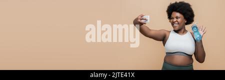 Happy african american plus size Frau hält Sportflasche und winkende Hand während Videoanruf isoliert auf beige, Banner, Stock Bild Stockfoto