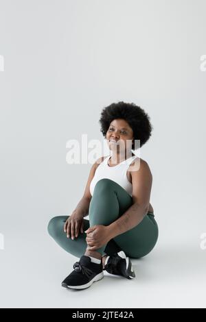 Durchdachte afroamerikanische Plus-Size-Frau in Sportswear und Sneakers auf grauem, Stock-Bild Stockfoto