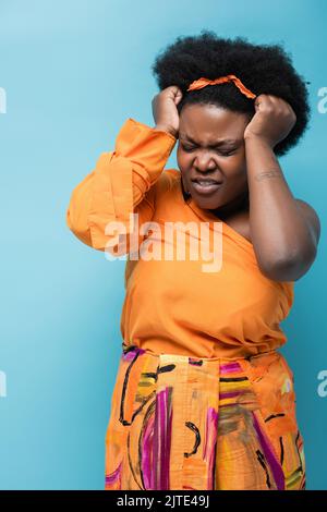 Lockige afroamerikanische Körper positive Frau in orange Kleid und Reifen Ohrringe mit Kopfschmerzen isoliert auf blau, Stock Bild Stockfoto