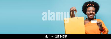 Fröhliche afroamerikanische Körper positive Frau hält Einkaufstasche und Kreditkarte isoliert auf blau, Banner, Stock Bild Stockfoto