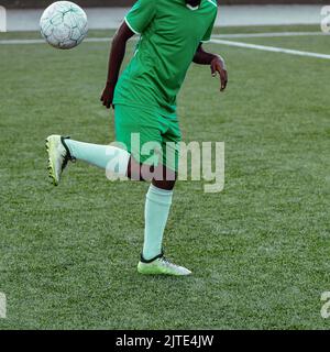 Nicht erkennbarer dunkelhäutiger, farbiger Mann in einer grünen Fußballuniform, der einen Fußballball hält, während er auf einem Fußballfeld qualifizierte Tricks macht Stockfoto