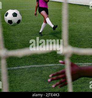 Eine nicht erkennbare dunkelhäutige Frau in einer pinkfarbenen Fußballuniform, die einen Fußball auf dem Fußballfeld beim Tortritt spielt. Hinter dem Tor. Stockfoto