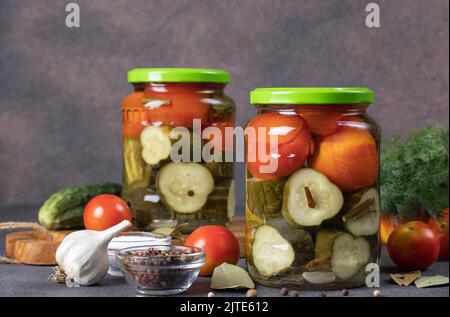 Eingelegte Gurken mit Kirschtomaten in zwei Gläsern auf braunem Hintergrund mit frischen Zutaten, Vorderansicht Stockfoto