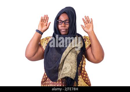 Ernsthafte muslimische reife Frau mit geschlossenen Augen, die die Hände hochhebt und betet. Stockfoto
