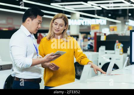 Asiatischer Berater Verkäufer im Geschäft für Elektronik und Haushaltsgeräte, verkauft eine Arbeitsmaschine an eine Frau, empfiehlt und genehmigt die Wahl Stockfoto
