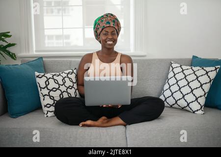 Lächelnde junge afroamerikanische Frau, die zu Hause mit gekreuzten Beinen auf ihrem Sofa sitzt und in die Kamera schaut Stockfoto