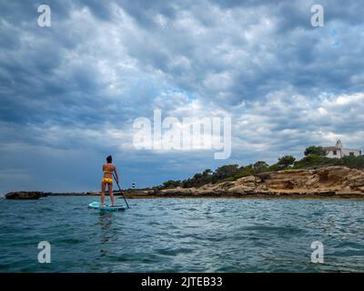 Frau, die auf einem Surfbrett unter einem dramatischen Himmel paddelt, Estalella, Küste von Llucmajor, Mallorca, Balearen, Spanien Stockfoto