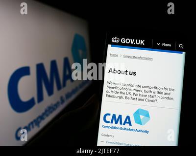 Person mit Mobiltelefon und Webseite der British Competition and Markets Authority (CMA) auf dem Bildschirm mit Logo. Konzentrieren Sie sich auf die Mitte des Telefondisplays. Stockfoto