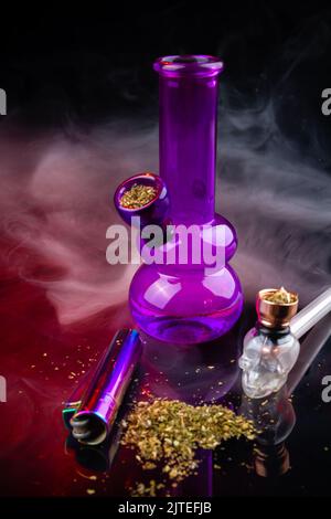 Vertikale Aufnahme von Glasbong mit Rauchrohr und Unkrauthaufen. Set von Marihuana und Rauchgeräten. Stockfoto