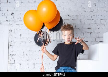 Halloween Kinder. Ein kleiner Junge in einem schwarzen T-Shirt mit orangefarbenen und schwarzen Ballons sitzt auf der Treppe des Hauses. Das Kind macht einen Schrecken Stockfoto