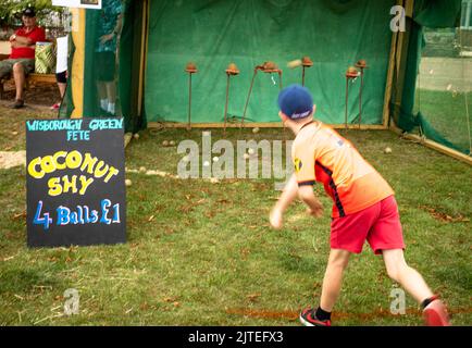 Ein kleiner Junge wirft eine Holzkugel auf Kokosnüsse auf einen traditionellen Kokosnussschüchtern im Wisborough Green Village Fete in West Sussex, Großbritannien. Stockfoto