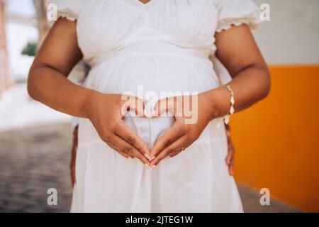 Eine Schwangere zeigt sich herzförmig am Bauch mit ihrem Partner dahinter Stockfoto