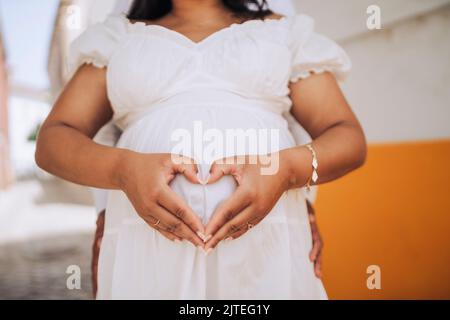 Eine Schwangere zeigt sich herzförmig am Bauch mit ihrem Partner dahinter Stockfoto