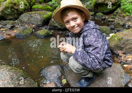 Junge in Gummistiefeln, hockt ein Hut eines Reisenden in der Nähe des Waldstroms Stockfoto