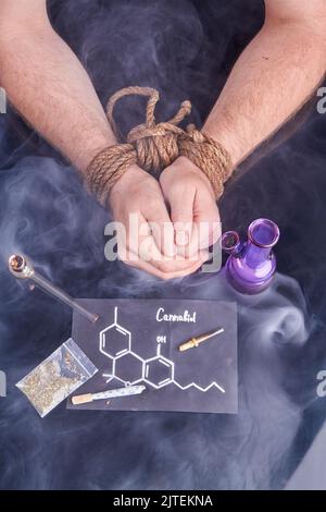 Sklave des Cannabiskonzepts. Gebundene männliche Hände mit Bong und Rauchpfeife. Molekül aus Cannabiol. Vertikale Aufnahme von oben. Stockfoto