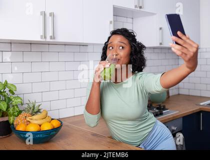 Eine junge multirassische Frau macht ein Selfie mit ihrem grünen Smoothie in der Küche Stockfoto