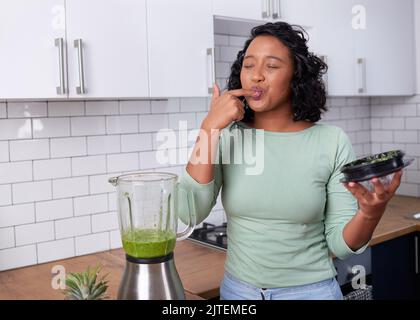 Eine junge, multiethnische Frau schmeckt ihren frisch zubereiteten grünen Smoothie in der Küche Stockfoto