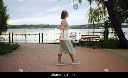 Eine kaukasische Frau, die mit Kopfhörern durch den Park im Hintergrund des Sees läuft Stockfoto