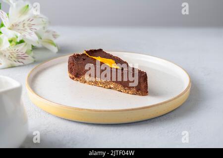 Scheibe roher veganer Schokoladenkuchen auf weißem Teller mit Blumen auf dem Hintergrund. Gesundes veganes Dessert, ohne Backen, ohne Eier, ohne Zucker Stockfoto