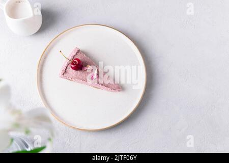 Draufsicht auf eine Scheibe roher Kirschkuchen auf weißem Teller auf neutralem grauen Hintergrund. Gesundes, rohes Dessert Stockfoto