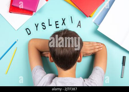Traurig müde frustriert Junge liegt auf dem Tisch mit vielen Büchern. Wort Dyslexie auf hellblauem Hintergrund. Legasthenie, Lernbehinderung, Lesen Stockfoto