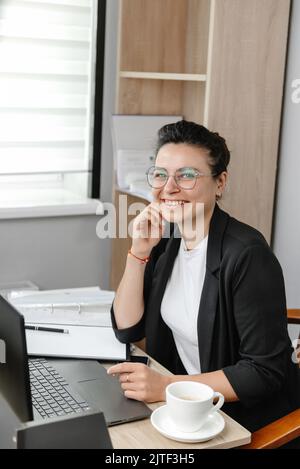 Eine junge Frau, die in einem echten Büro eine Brille trägt, lächelt und schaut auf die Kamera. Vertikales Foto. Hybrid-Arbeit. Kleines Büro. Stockfoto