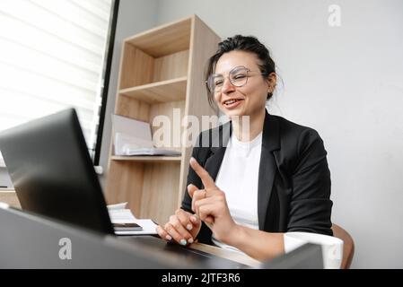 Das Mädchen im Büro führt eine Online-Konferenz durch und sagt etwas. Arbeiten in einem Büro-Hybrid-Arbeitssystem Stockfoto