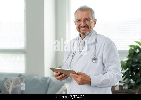 Porträt eines älteren Arztes, er schaut in die Kamera. Der Arzt steht vor dem Hintergrund einer medizinischen Klinik und hält eine Tablette in den Händen. Stockfoto