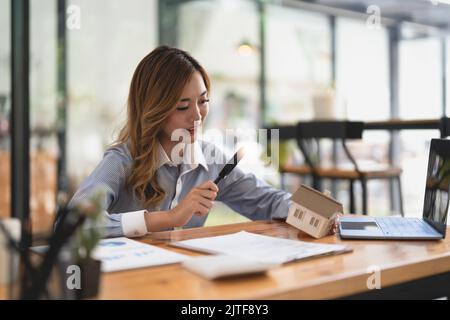 Auf der Suche nach Immobilienagentur, Immobilienversicherung, Hypothekendarlehen oder neues Haus. Frau mit Lupe über einem Holzhaus in ihrem Büro Stockfoto